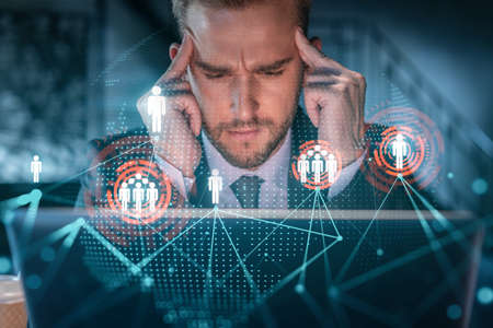 Handsome businessman in suit at workplace working with laptop to hire new employees for international business consulting. HR, social media hologram icons over office backgroundの写真素材
