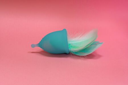 Blue Menstrual cup with blue feathers on pink background. Alternative feminine hygiene product...