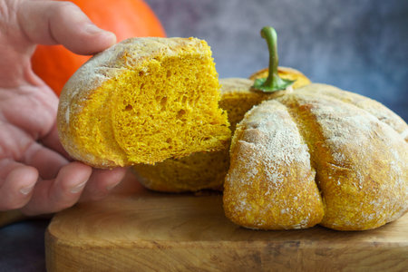 Close-up Cutaway Homemade sourdough bread in the shape of a pumpkin on a gray background. A man's hand holds a piece of pumpkin bread. Autumn pumpkin bun.の写真素材