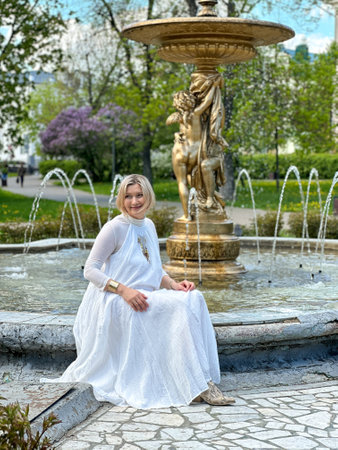 urban portrait of a middle-aged woman against the background of a fountain. Girl Dressed in a white sundress.の写真素材