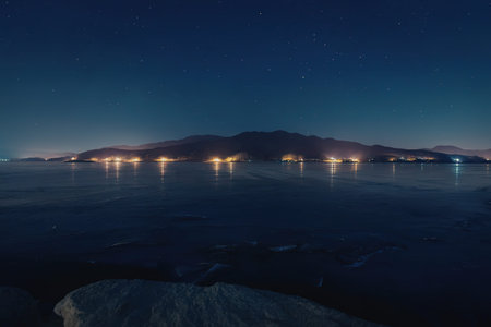 A serene nighttime scene featuring a mountain range across a body of water with lights reflecting on its surfaceの写真素材