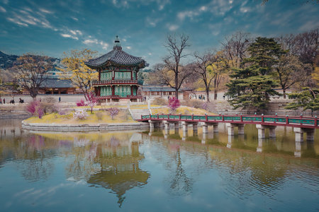A tranquil pond scene featuring a traditional Asian-style pavilion surrounded by trees and vibrant flowers, with a bridge extending into the water.の写真素材