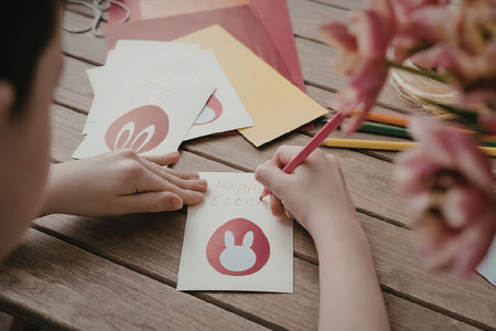 A person's hands are shown coloring on a piece of paper with a red pencil, surrounded by various art...