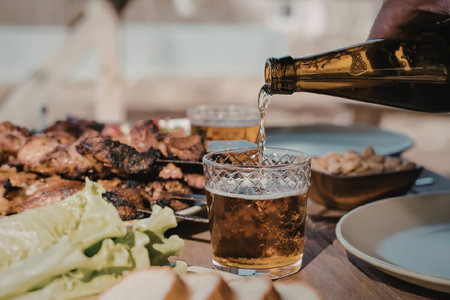 A hand pours a glass of beer from a bottle, with a table full of food and drinks in the background.