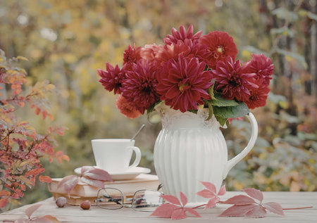 A white vase with red flowers, a cup and saucer, and fallen leaves on a table outdoors in an autumn setting.