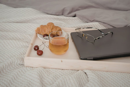 A breakfast setup on a bed with a croissant, a glass of juice, and a laptop