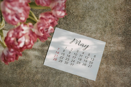 A calendar page for the month of May, placed on a green surface with pink flowers in the top left...