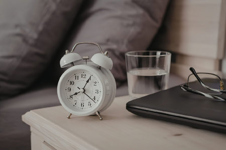 A still life of a bedside table with an alarm clock, a glass of water, a laptop, and glasses.