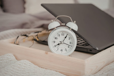 An alarm clock, glasses, and a laptop on a wooden tray on a bed