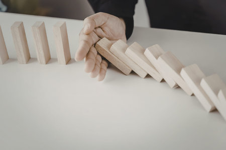 A hand stopping a row of falling wooden dominoes on a white table.の写真素材