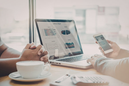 Two people sitting at a table with a laptop and smartphone, with a cup of coffee and notebook...