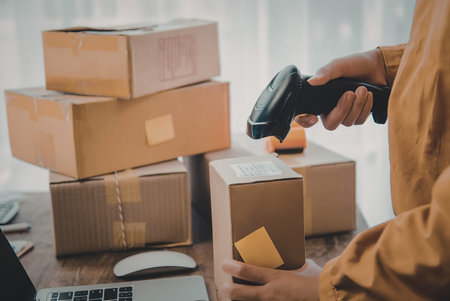 A person in an orange shirt scans a cardboard box with a barcode scanner, surrounded by stacked...