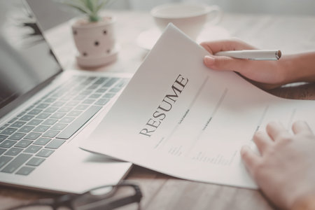 A person is holding a resume with a pen, sitting next to a laptop on a wooden desk.の写真素材