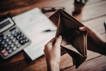 A person holding an empty brown leather wallet with a calculator, papers, and pen on a wooden desk.