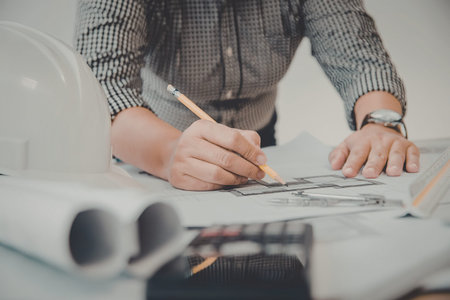 A man in a gray shirt working on a blueprint with a pencil, surrounded by rolled up papers and a...