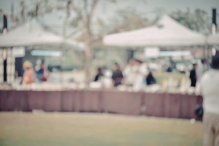 A blurry image of people standing under white tents in a park with brown barrier and green grass