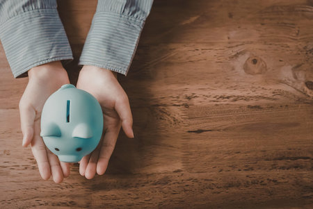 A person holding a light blue piggy bank in their hands on a wooden table, emphasizing saving and finance.の写真素材