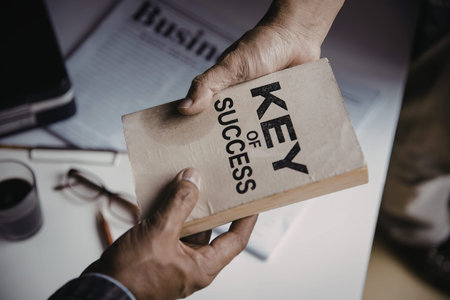 Two hands holding a book with the words 'Key to Success' printed on it, placed on a desk with office...