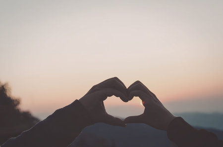 A romantic gesture of two hands forming a heart shape against a beautiful sunset background.の写真素材