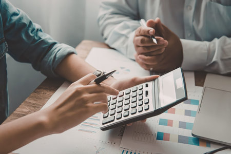Two individuals sitting at a desk, engaged in a discussion while looking at financial data and using...