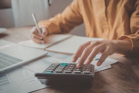 A person in an orange shirt using a calculator and writing on a notebook with a pen, surrounded by...