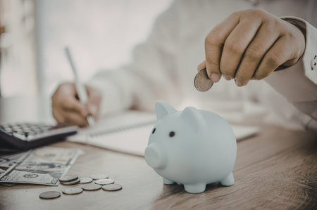 A person is putting coins into a white piggy bank on a desk with a calculator and papers.の写真素材