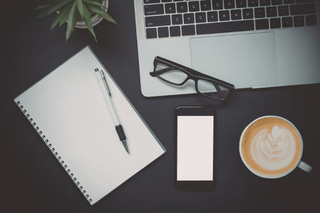 A flat lay of a modern workspace featuring a laptop, notebook, phone, coffee, and glasses on a black...