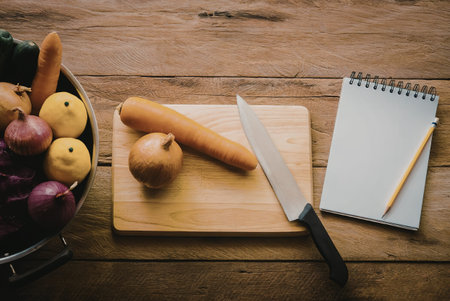 A wooden table with a bowl of fresh vegetables, a cutting board with a carrot and onion, a knife, and a notepad with a pencil.の写真素材