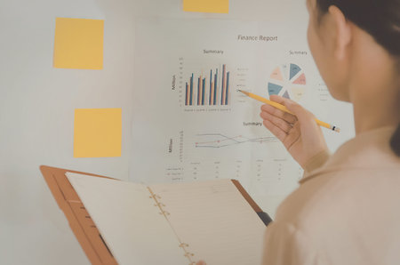 A woman in a beige shirt is analyzing data on a wall chart with colorful graphs and sticky notes.の写真素材