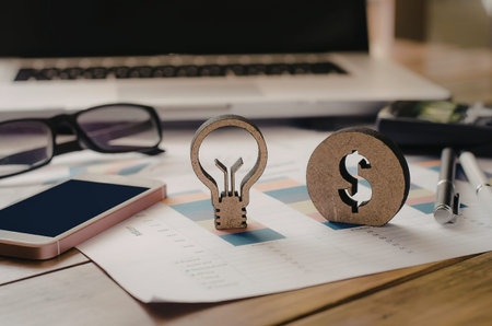 A creative business concept featuring a light bulb and dollar coin on a desk with a laptop, phone, and glasses in the background.の写真素材