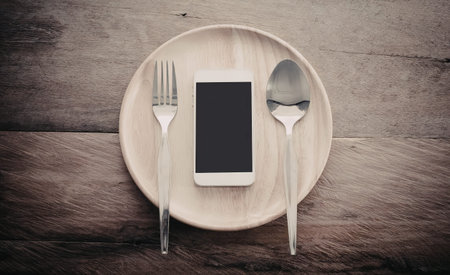 A smartphone placed on a wooden plate with a fork and spoon on a wooden table