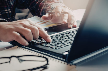 A person typing on a laptop keyboard with a notebook and glasses on the desk