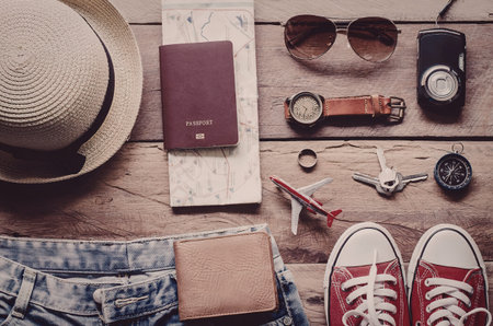 A collection of travel essentials including a hat, passport, wallet, sunglasses, watch, camera, and clothing laid out on a wooden surface.の写真素材