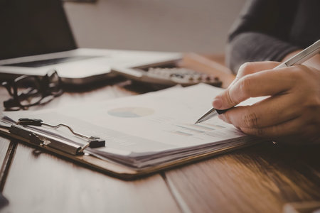 A person is sitting at a desk, holding a pen and filling out a document on a clipboard. The...