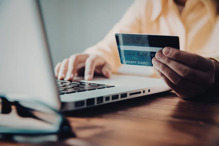 A person is sitting at a desk, holding a credit card in one hand and typing on a laptop with the...