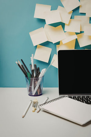 A white desk with a black laptop, a spiral notebook, a purple cup of pens and pencils, and a wall...