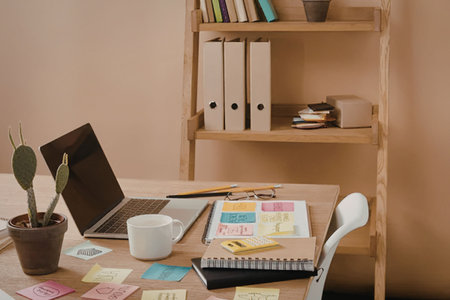 A wooden desk with a laptop, notebook, coffee mug, and colorful sticky notes, with a bookshelf in...