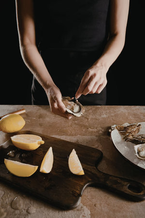 a person wearing a black shirt standing behind a table, opening an oyster with a knife, with a...