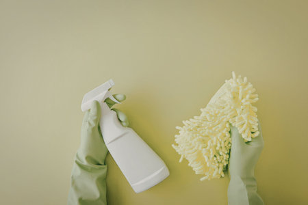A hand in a green glove holding a white spray bottle and another hand holding a green microfiber...