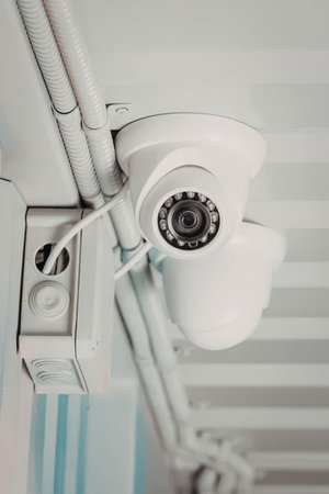 A white security camera is mounted on a wall, with cables and pipes visible in the background. The...