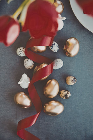 A collection of gold and speckled Easter eggs, red tulips, and a red ribbon on a gray surface.
