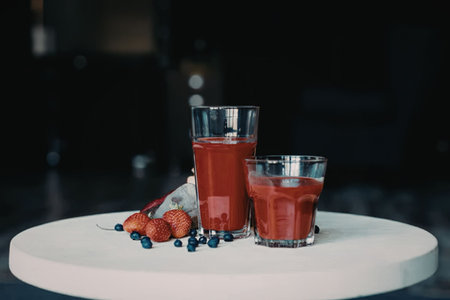Two glasses of red juice on a white table with some blueberries and strawberries