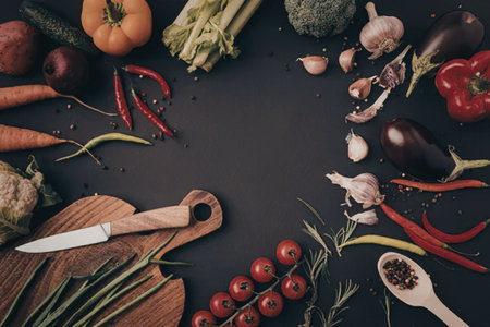 An assortment of fresh vegetables and herbs arranged on a black surface with a knife and cutting...