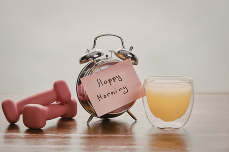 A still life of a morning routine setup featuring dumbbells, an alarm clock with a 'Happy Morning' note, and a glass of orange juice on a wooden surface.の写真素材