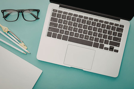 A laptop, glasses, pens, and a notepad on a teal desk