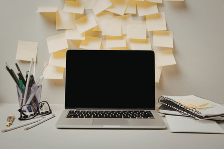 A laptop sits on a cluttered desk with post it notes, pens, and notebooks.