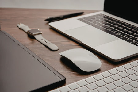 A modern workspace setup featuring a laptop, keyboard, mouse, smartwatch, and tablet on a wooden...