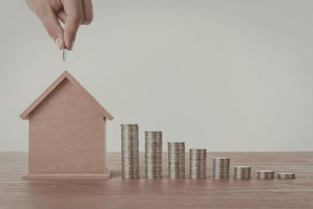 A hand putting a coin into a miniature house next to stacks of coins