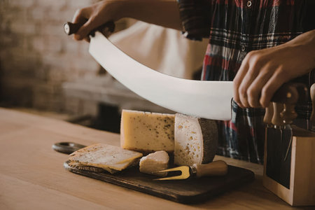 A woman in a plaid shirt cutting a large piece of cheese on a wooden table with a knife.