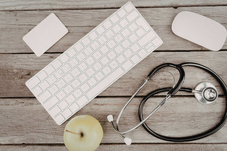 A stethoscope, keyboard, mouse, and green apple on a wooden deskの写真素材